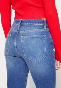 Vaqueros de denim azul de corte recto y tiro alto, con dos bolsillos traseros y un ligero detalle de desgaste. Combinados con una blusa roja de canalé.