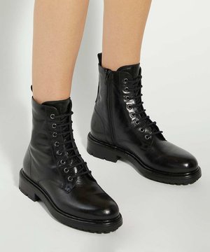Bottines noires en cuir à lacets avec fermetures éclair latérales portées sur jambes nues sur un fond clair uni.