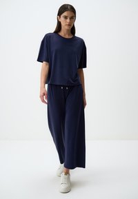Completo da casa blu navy composto da una maglietta corta e ampia a maniche corte e pantaloni a gamba larga. Realizzato in un tessuto morbido con un leggero lucido, impreziosito da dettagli in cordoncino.