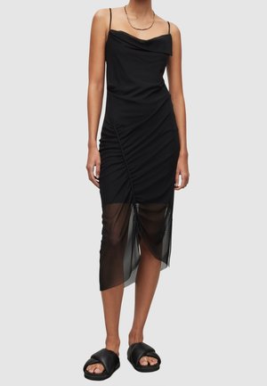 Femme portant une robe noire sans manches froncée avec un ourlet asymétrique transparent et des sandales noires à enfiler, debout devant un fond uni.