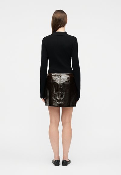 Γυναίκα που στέκεται με την πλάτη προς τα εμπρός, φορώντας μαύρο μακρυμάνικο τοπ, γυαλιστερή μαύρη δερμάτινη μίνι φούστα και μαύρα παπούτσια χωρίς κορδόνια, σε λευκό φόντο.