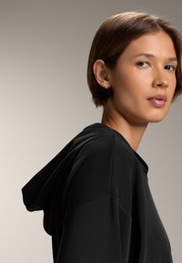 Felpa nera con cappuccio realizzata in tessuto morbido, con cappuccio regolabile, vestibilità comoda e cuciture sulle spalle con leggere pieghe per aggiungere texture.