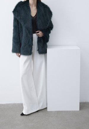 Personne portant une veste en fausse fourrure vert foncé, un top noir et un pantalon blanc à jambes larges, debout à côté d’un piédestal rectangulaire blanc sur un sol gris.