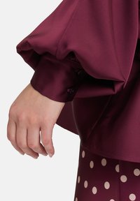 Burgundy satin blouse with voluminous sleeves, buttoned cuffs, and a smooth texture, paired with polka dot-patterned fabric on the bottom.