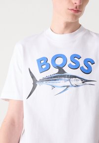 Camiseta de algodón blanca con un gráfico de un marlín azul y negro y la palabra "BOSS" en grandes letras azules arriba. Manga corta, cuello redondo.