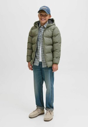 Veste verte matelassée avec une capuche, superposée sur une chemise à carreaux et un t-shirt blanc. Portée avec un jean bleu clair et des chaussures en daim grises. Casquette en denim.