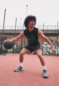 Schwarzes ärmelloses Shirt mit grünem und pinkem Basketball-Graphic, gemusterte Shorts mit rotem Saum, graue Sportschuhe, dribbelt einen Basketball auf einem Platz.