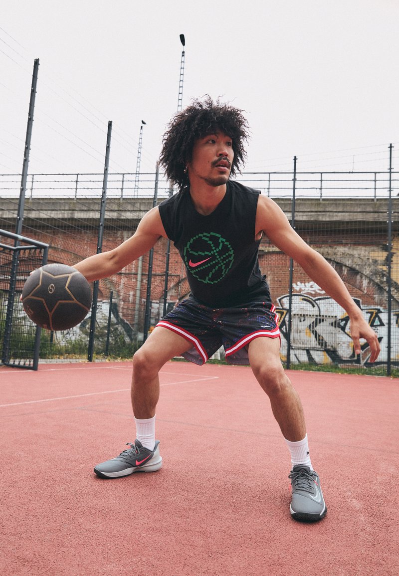 T-shirt sans manches noir avec graphique de basket-ball vert et rose, short à motifs avec bord rouge, chaussures de sport grises, en train de dribbler un ballon sur un terrain.