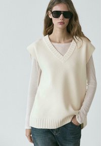 Sweatervest i cremefarvet strik med V-udskæring og ribbet kant; lagdelt over en let langærmet top. Båret med mørke denimjeans.