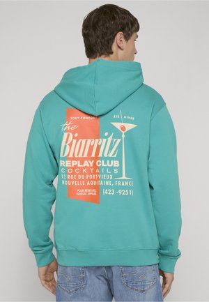 Mand iført en teal hættetrøje med orange og hvid grafisk tekst om Biarritz-cocktails og et martiniglas på ryggen.