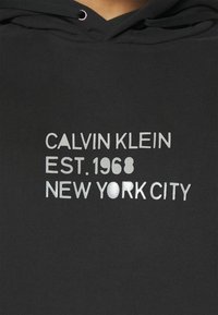 Felpa nera con cappuccio con testo stampato argento: "CALVIN KLEIN EST. 1968 NEW YORK CITY," con una texture a coste e cappuccio con cordino.