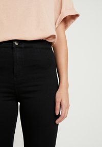 Svart högmidjade jeans med en slät yta och ton-i-ton sömmar, med en framknäppning med knapp och dragkedja; parat med en ljusrosa oversized topp.