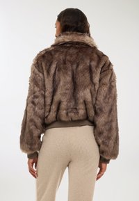 Giacca corta in finta pelliccia marrone con colletto alto e polsini a costine, caratterizzata da una superficie testurizzata e un aspetto morbido.