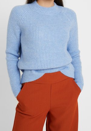 Frau trägt einen hellblauen Strickpullover, der in rostfarbenen, hoch taillierten Hosen mit den Händen in den Taschen steckt.
