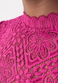 Gros plan d'une personne portant un haut en dentelle rose vif avec des motifs floraux et un col haut festonné.