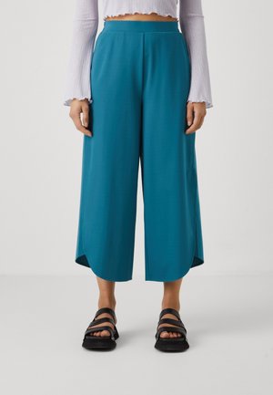 Mujer con pantalones culotte anchos de color verde azulado, top corto de manga larga blanco y sandalias negras, de pie sobre un fondo liso.