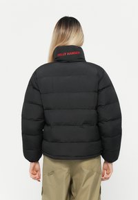 Chaqueta de plumas negra con secciones cosidas horizontalmente. Presenta un cuello alto y el logo rojo "HELLEY HANSEN" en la parte posterior. Material resistente.