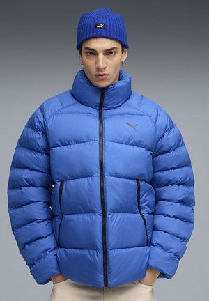 Jonge man in een blauwe Puma pufferjack en bijpassende beanie, staand met zijn handen in de zakken tegen een grijze achtergrond.