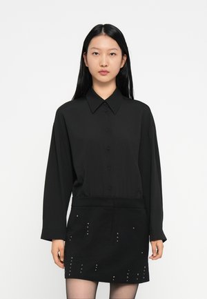 Donna che indossa una camicia nera a maniche lunghe con bottoni e una gonna nera con piccoli chiodi metallici, in piedi contro uno sfondo bianco semplice.