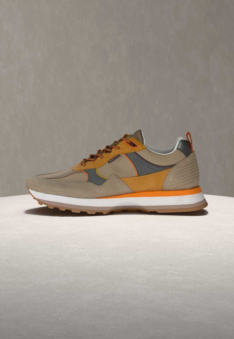 Αθλητικό παπούτσι σε μπεζ και πορτοκαλί χρώμα με λευκή σόλα, γκρι και κίτρινες λεπτομέρειες και υφασμάτινη υφή, τοποθετημένο σε ανοιχτή επιφάνεια με φόντο γκρι.