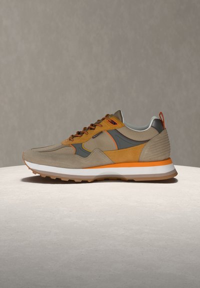 Béžovo-oranžový sportovní teniska s bílou podrážkou, šedými a žlutými akcenty a texturovanou tkaninou, vystavená na světlém povrchu proti šedému pozadí.