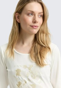Blonde Frau mit blauen Augen schaut nach rechts, trägt ein weißes Oberteil mit einem subtilen beigen abstrakten Muster auf der Vorderseite, vor einem hellgrauen Hintergrund.