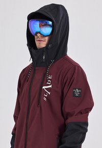 Bordeaux ski-jack met een zwarte capuchon, voorzien van een voorgesluiter, koordjes en een logo patch op de mouw. Reflecterende blauwe skibril wordt gedragen.