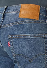 Vaqueros de mezclilla en azul oscuro con acabado texturizado. Presentan un parche de cuero marrón en la pretina, etiqueta roja de Levi's y costuras amarillas en contraste.