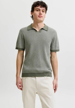 Młody mężczyzna w krótkim rękawie, teksturowanej szaro-zielonej koszulce polo i kremowych spodniach, stoi na tle jednolitego jasnego tła.