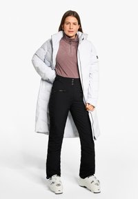 Lange weiße Daunenjacke, helllila Fleece-Oberteil, schwarze Skihosen mit Reißverschlüssen und weiße Skischuhe mit Akzenten. Steht vor einem weißen Hintergrund.