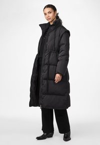 Svart dunjakke med høy krage, oversized design, quiltete paneler og glidelås. Båret over et svart antrekk med høye støvler.