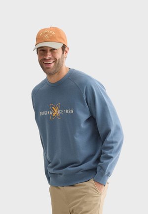 Sudadera azul claro con puños acanalados, que presenta el texto naranja "ORIGINAL SINCE 1939" y un gráfico. Combinada con una gorra beige y naranja.
