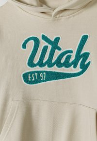 Beige színű kapucnis pulóver, melyen egy teal színű, kiemelkedő textúrájú, hímzett "Utah" logó található, valamint "EST 97" felirat krém színben, középen a zsebből.