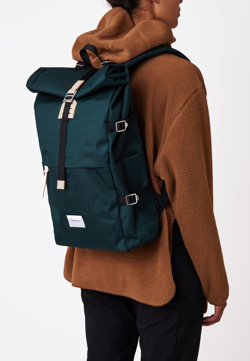 Personne portant une veste polaire marron et un pantalon noir avec un grand sac à dos à rabat roulé vert foncé, sécurisé par des sangles noires et des boucles beige.
