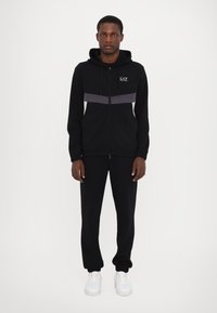 Black zip-up hoodie with gray and white horizontal stripes, drawstring hood, paired with black joggers and white sneakers.