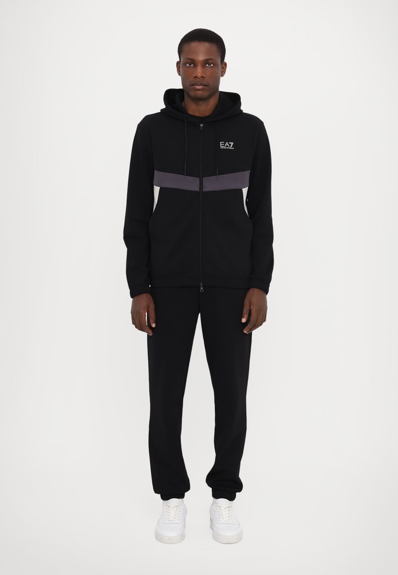Black zip-up hoodie with gray and white horizontal stripes, drawstring hood, paired with black joggers and white sneakers.