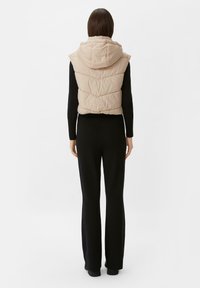 Beżowa kamizelka puffer z kapturem, z wzorem w chevronowy quilt. Nosi na sobie czarną bluzkę z długim rękawem i czarne spodnie, widok z tyłu.