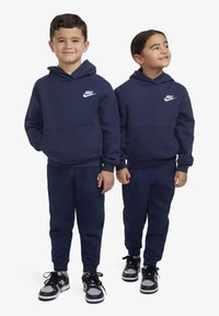 Sweatshirts com capuz azul-marinho, com um bolso frontal e logótipos brancos da Nike, combinados com calças escuras e ténis pretos.