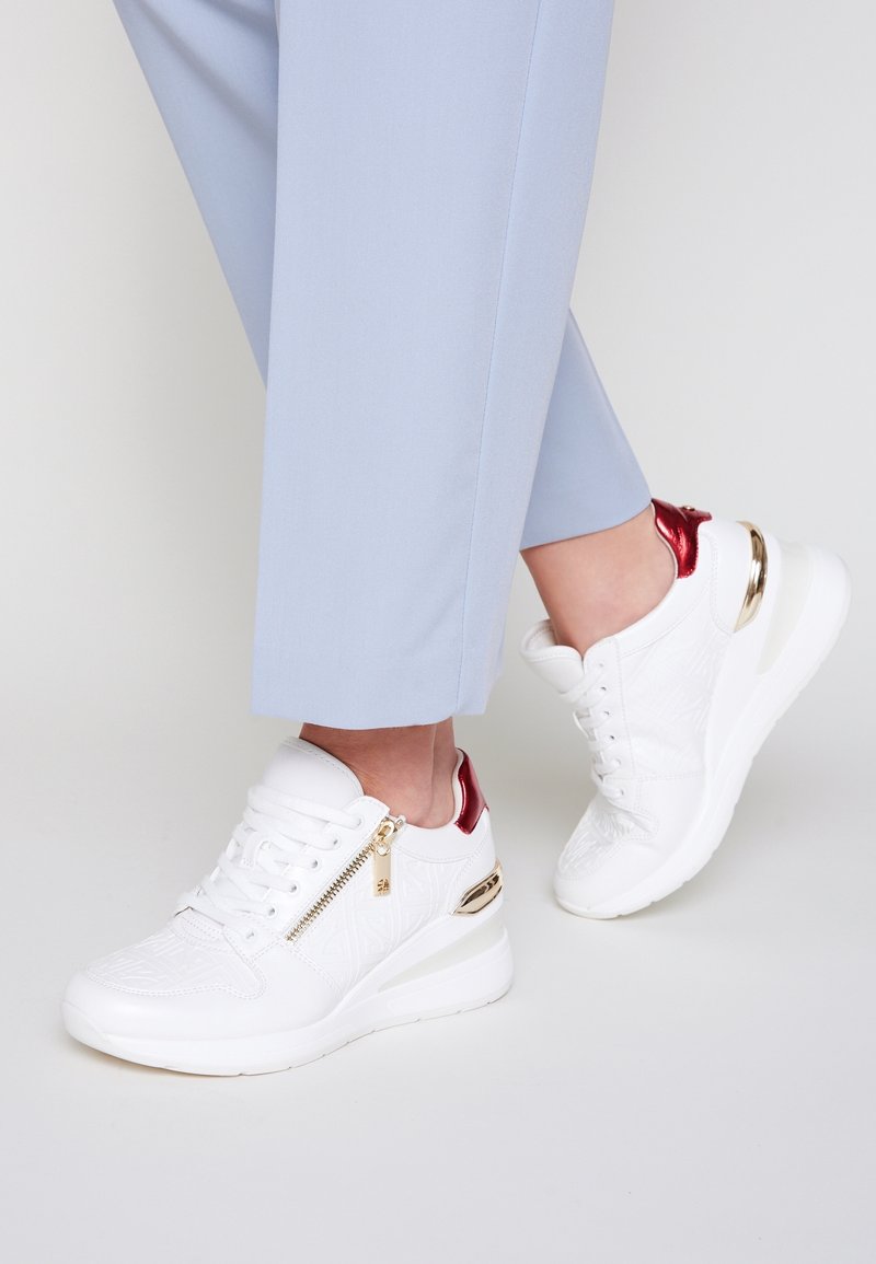 Chaussures de sport blanches avec un motif texturé, des accents rouges, des éléments dorés et une semelle compensée, portées avec un pantalon court bleu clair.