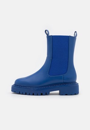 Bota Chelsea de cuero azul con paneles laterales elásticos, tiradores y suela robusta con dibujo, mostrada sobre un fondo blanco.