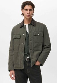 Giovane uomo che indossa una giacca verde oliva con bottoni, con tasche a patta e colletto scuro, sovrapposta a un gilet in maglia e una camicia bianca.
