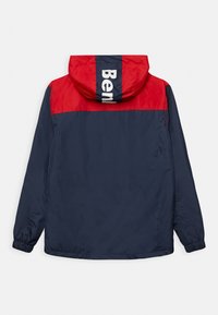 Veste coupe-vent bleu marine et rouge avec capuche, arborant le logo blanc "Belt" au dos, des poignets élastiques et une texture lisse.