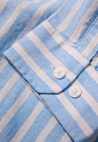 Colletto di camicia in cotone a righe azzurre e bianche con due bottoni bianchi, caratterizzato da una texture liscia e dettagli cuciti.