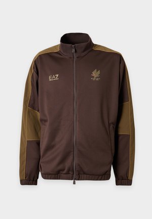 Homme debout portant une veste zippée marron, une chemise blanche, un pantalon noir et des baskets noires sur un fond clair uni.