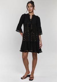 Abito nero realizzato in tessuto testurizzato con maniche a campana, rifiniture in pizzo e accenti dorati. Presenta un design a strati con pendagli e motivi circolari.