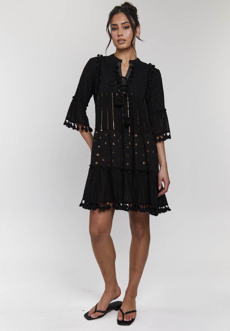 Abito nero realizzato in tessuto testurizzato con maniche a campana, rifiniture in pizzo e accenti dorati. Presenta un design a strati con pendagli e motivi circolari.