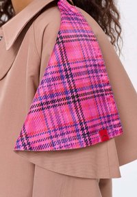 Beżowy płaszcz z klapą na ramieniu, w której znajduje się panel z materiału w kratę w kolorach różowym, fioletowym i czarnym, z małą czerwoną metką.