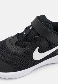 Černá sportovní bota s texturovaným síťovaným svrškem, gumovou podrážkou, bílým logem Nike swoosh a černým páskem přes střední část chodidla.