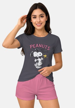 Woman with long dark hair wearing a dark gray Peanuts T-shirt with Snoopy and Woodstock and pink shorts, smiling against a plain background.