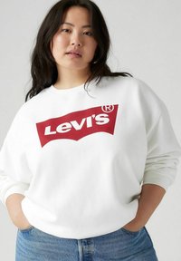 Felpa bianca con un grande logo rosso di Levi's. Presenta un taglio rilassato e un collo a giro. Abbinata a jeans in denim blu chiaro.